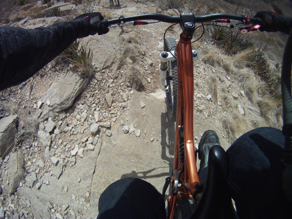 Mountain biking perspective from above the handlebars, showing a rocky trail ahead. The rider's hands gripping the handlebars and parts of their jeans and shoes are visible. Surrounding terrain features sparse vegetation and rough, rocky ground. Roughrider Canyon Freeride Park mountain bike trail.