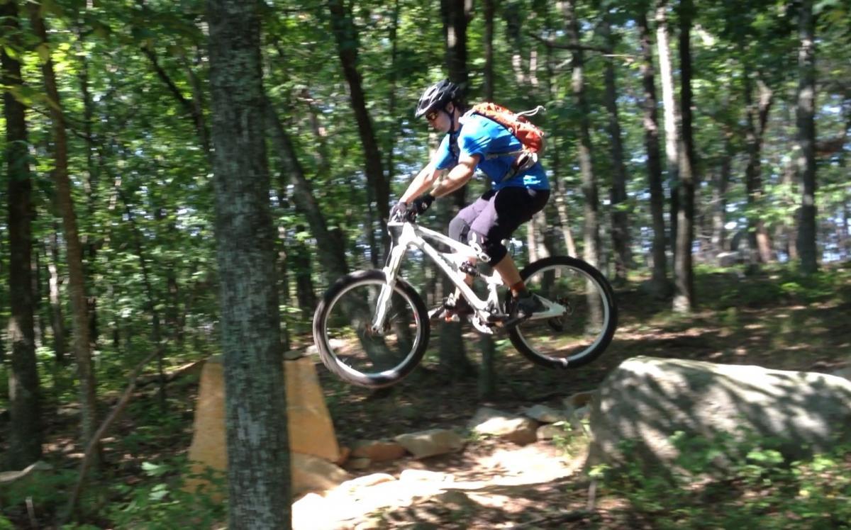 A cyclist in a blue shirt and black shorts jumps off a rock while mountain biking through a wooded trail. The scene features dense greenery and trees in the background, showcasing an adventurous outdoor environment. Vietnam Trails mountain bike trail.