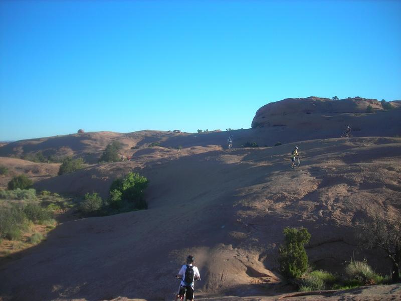 A scenic outdoor landscape featuring several mountain bikers riding across rocky terrain under a clear blue sky. The background showcases rolling hills and sparse vegetation, with a few small bushes and trees in the foreground. Slickrock mountain bike trail.