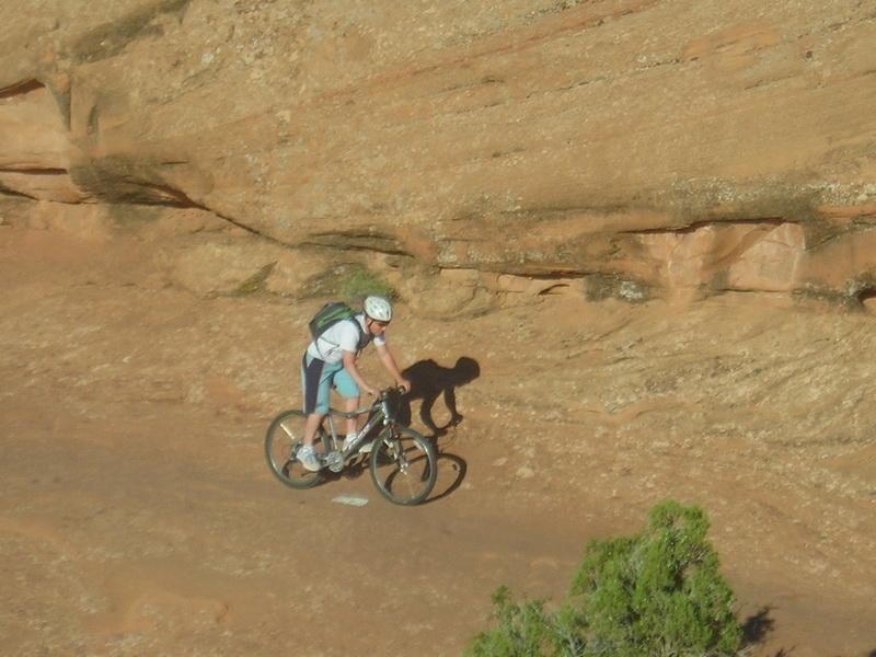 A person biking on a rocky trail surrounded by red sandstone formations, with a green shrub in the foreground. The cyclist is wearing a helmet and a backpack, navigating the terrain under bright sunlight. Slickrock mountain bike trail.