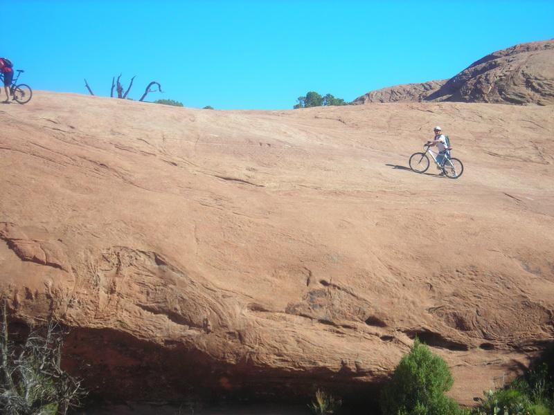 A cyclist riding on a smooth, reddish rock surface under a clear blue sky, with another cyclist visible in the background. The landscape features natural rock formations and sparse vegetation. Slickrock mountain bike trail.