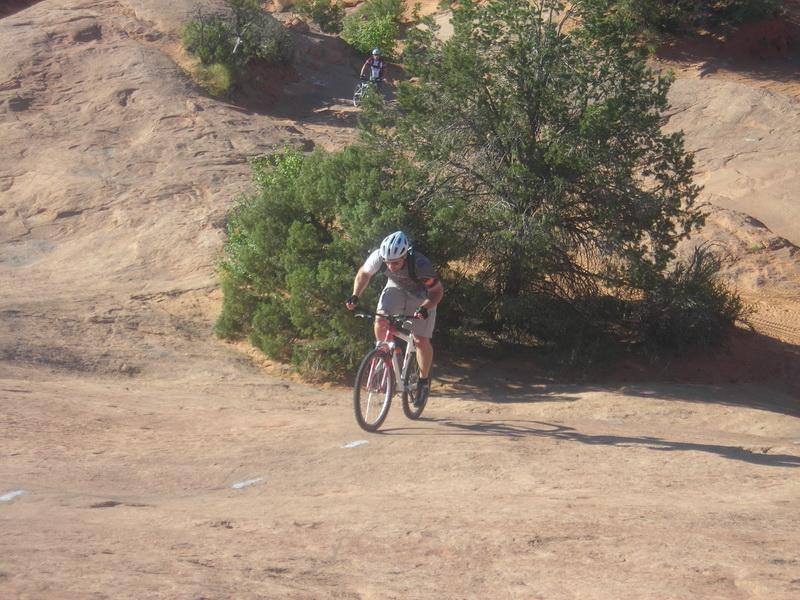 A person riding a mountain bike uphill on rocky terrain, with greenery and another cyclist visible in the background. Slickrock mountain bike trail.