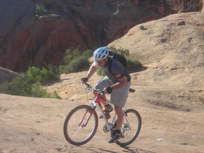 A person riding a mountain bike on a rocky terrain, wearing a helmet and a backpack, focused on navigating the uneven surface in a natural outdoor setting with greenery in the background. Slickrock mountain bike trail.