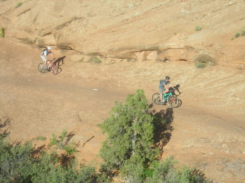 Two mountain bikers navigate a rugged trail along rocky terrain. One rider is dressed in a white and black outfit, while the other wears a dark top and has a backpack. A small tree and some shrubs are visible near the path, surrounded by the dry, rocky landscape. Slickrock mountain bike trail.