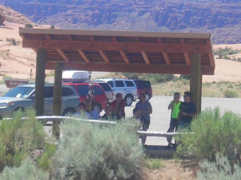 Group of people standing under a wooden shelter at a parking area, with vehicles nearby and a rocky landscape in the background. Green shrubs are in the foreground, and the scene appears to be in a desert or arid region. Slickrock mountain bike trail.