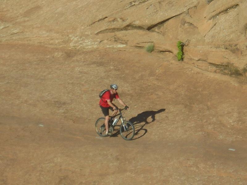 A cyclist in a red shirt rides a mountain bike on a rocky, sunlit trail. The terrain appears rugged and dry, with sparse vegetation and a clear blue sky in the background. Slickrock mountain bike trail.