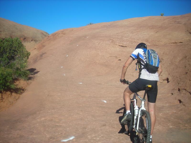 A mountain biker riding uphill on a rocky terrain under a clear blue sky, with a backpack visible on their back. The landscape features a natural rocky surface with scattered greenery on the left. Slickrock mountain bike trail.