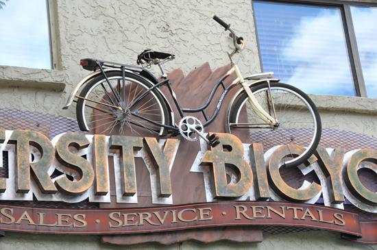 A vintage bicycle mounted on a sign for "University Bicycle," which includes the words "Sales," "Service," and "Rentals." The bike is positioned above the business name against a textured wall with windows in the background.