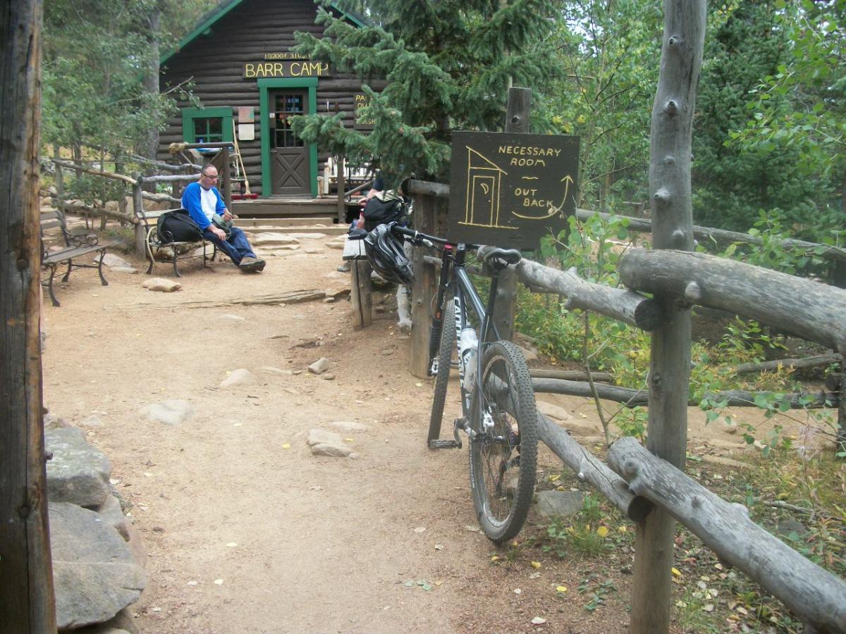 A rustic scene at Barr Camp featuring a log cabin in the background, with a sign indicating the location of a restroom. A mountain bike is leaning against a wooden fence in the foreground, while a man sits on a bench nearby, enjoying the surroundings. The area is surrounded by trees and a dirt pathway, creating a tranquil outdoor atmosphere. Barr Trail / Pikes Peak mountain bike trail.