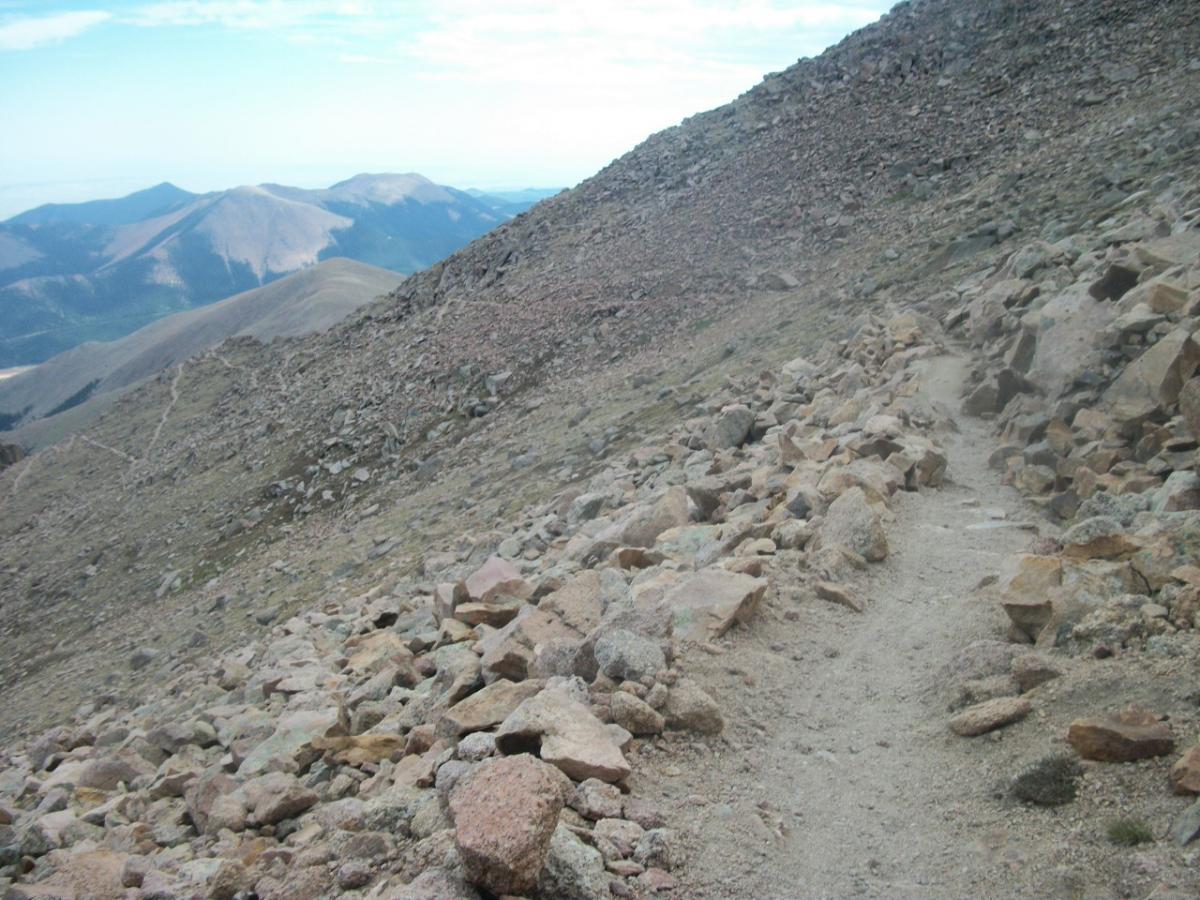 A rocky, uneven hiking trail winding along a mountain slope with scattered boulders, overlooking distant ridges and peaks under a partly cloudy sky. Barr Trail / Pikes Peak mountain bike trail.