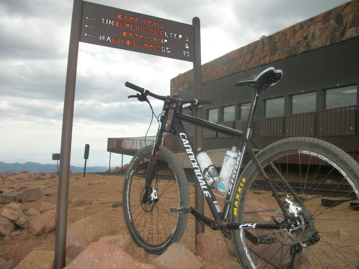 A mountain bike parked next to a trail sign marking the Barr Trail, Timberline Shelter, Barr Camp, and Manitou Springs distances. The background features rocky terrain and cloudy skies, with a building visible in the distance. Barr Trail / Pikes Peak mountain bike trail.