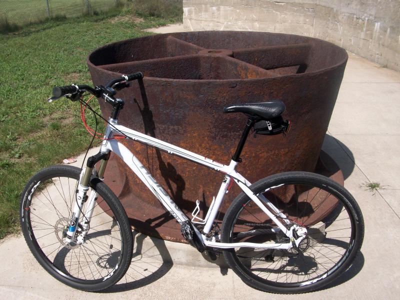 Raleigh Talus 29er: A white mountain bike is leaning against a large, rusty circular metal structure on a concrete surface, with green grass visible in the background. The structure appears to be an industrial piece, possibly a water tank or a similar object. The scene is illuminated by sunlight, highlighting the bike and the texture of the rust.