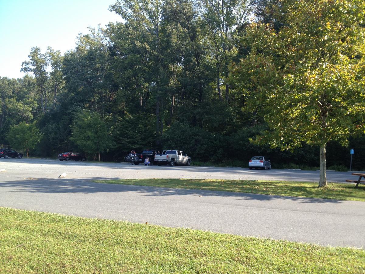 Parking lot surrounded by trees, with several vehicles parked on a sunny day. A person is seen near a truck, possibly unloading or loading items, and a few grassy patches are visible in the foreground. Walnut Creek mountain bike trail.