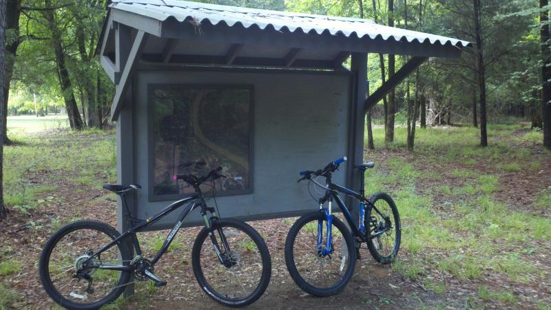 Specialized Myka Disc: Two mountain bikes are parked beside a covered information kiosk in a wooded area. The kiosk features a large map displayed on its front. Surrounding the bikes and kiosk is a lush green landscape, with trees in the background and fallen leaves scattered on the ground.