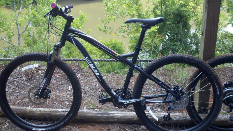 Specialized Myka Disc: A black mountain bike with thick tires and a sturdy frame is positioned next to a wooden railing overlooking a body of water. Lush greenery surrounds the area, adding a natural backdrop to the scene.
