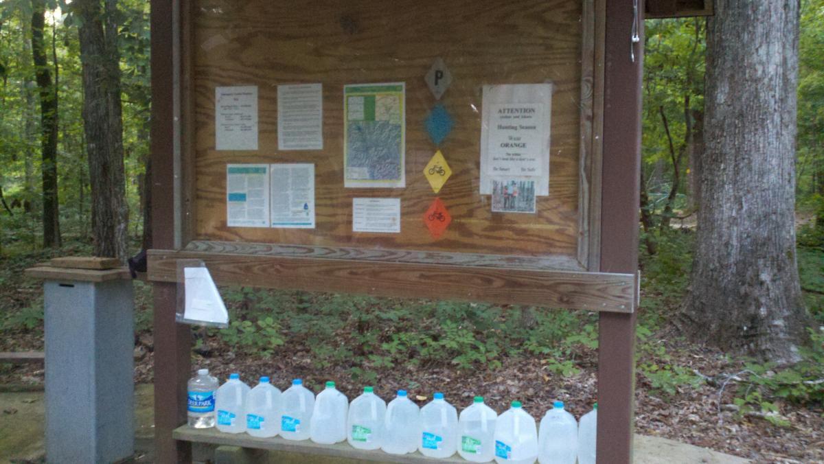 A bulletin board in a wooded area displaying various informational papers and signs, including a caution notice for hunting season, along with several plastic jugs of water lined up at the bottom. The surrounding environment features trees and greenery. Noxubee Crest mountain bike trail.
