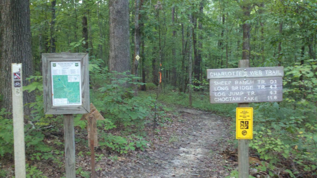 A wooded trail entrance sign indicating "Charlotte's Web Trail," with directional arrows and mileage markers to various locations including Sheep Ranch Trail, Long Bridge Trail, Log Jump Trail, and Choctaw Lake. A trail map is displayed on a nearby post, surrounded by lush green foliage and trees. Noxubee Crest mountain bike trail.