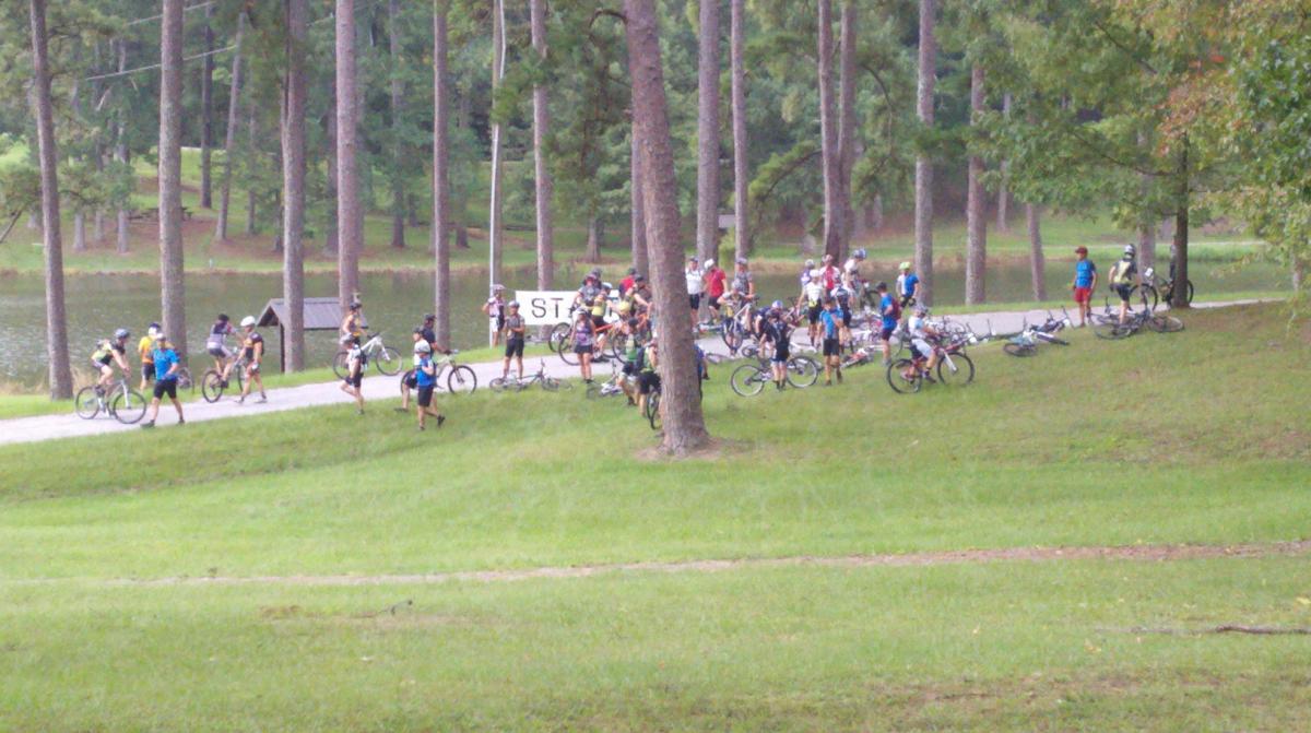 A group of cyclists gathered by a lake, with some riders on bicycles and others standing on the grass. There is a sign labeled "START" visible in the background, indicating the start of a cycling event. Tall trees surround the area, providing a natural backdrop. Noxubee Crest mountain bike trail.