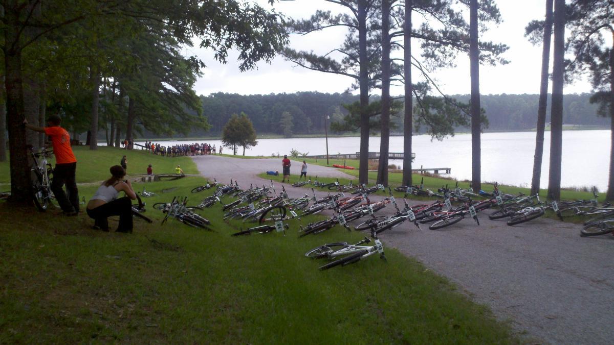 A group of bicycles scattered on a grassy area near a lake, with several people in casual clothing nearby, some sitting and others standing. Trees line the path leading to the water, where a small gathering of people can be seen in the background. Noxubee Crest mountain bike trail.
