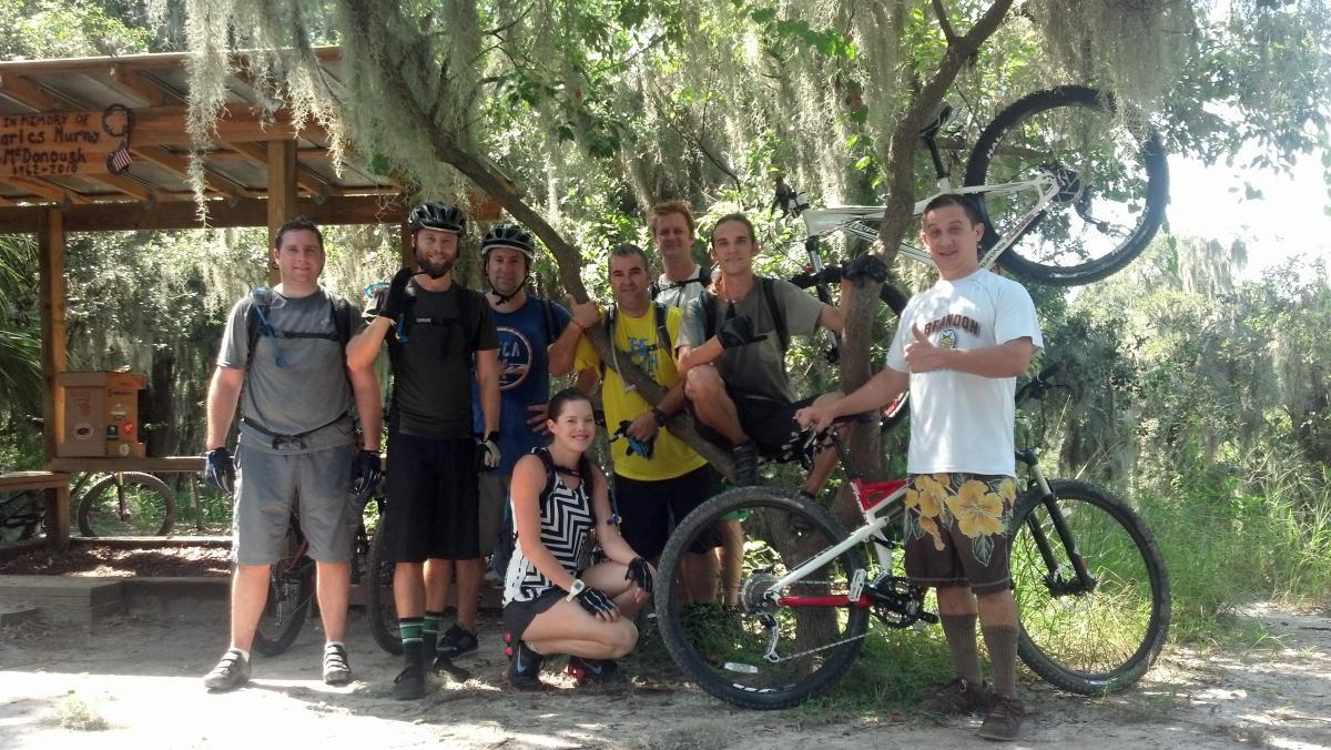 Group of nine mountain bikers posing together outdoors, surrounded by trees with Spanish moss. They are wearing biking gear and helmets, with several bicycles in the background, including one leaning against a tree. The scene conveys a sense of camaraderie and outdoor adventure. Loyce E. Harpe Park mountain bike trail.