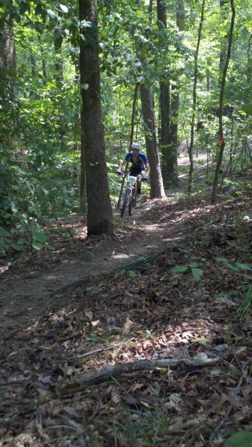 A person riding a mountain bike along a dirt trail in a wooded area, surrounded by tall trees and lush green foliage. The rider is wearing a blue jersey and a helmet, navigating through the natural terrain covered with fallen leaves and twigs. Noxubee Crest mountain bike trail.
