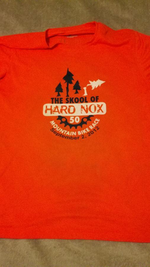 An orange t-shirt featuring the design for "The Skool of Hard Nox 50" mountain bike race, dated September 2, 2012. The design includes graphic elements such as trees and a bicycle gear motif. Noxubee Crest mountain bike trail.