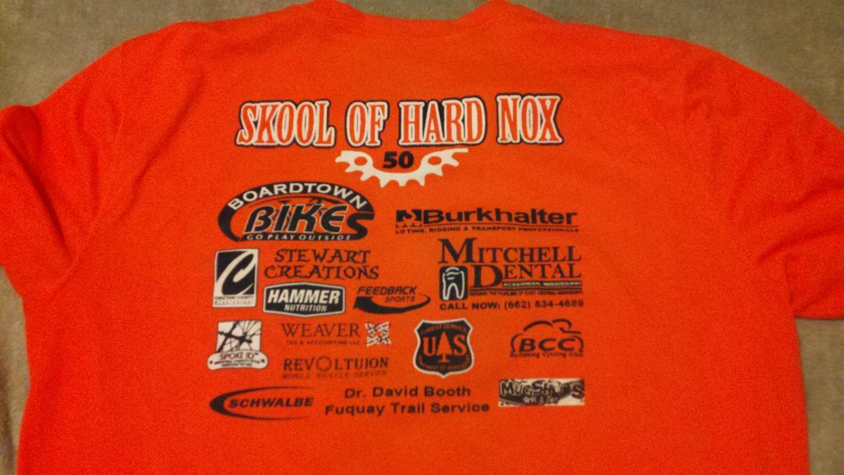 Image of the back of an orange t-shirt featuring the text "SKOOL OF HARD NOX" in bold lettering, accompanied by various sponsor logos, including Boardtown Bikes, Burkhalter, Stewart Creations, Hammer Nutrition, and others. The design also includes the number "50" and additional text related to the sponsors. Noxubee Crest mountain bike trail.