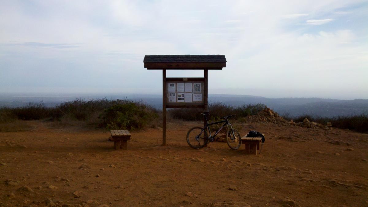 A scenic viewpoint at a trailhead featuring a wooden informational kiosk surrounded by barren land and sparse vegetation. A parked mountain bike leans against the kiosk, with two rustic wooden benches nearby. The horizon shows distant hills under a cloudy sky. Rancho La Costa mountain bike trail.