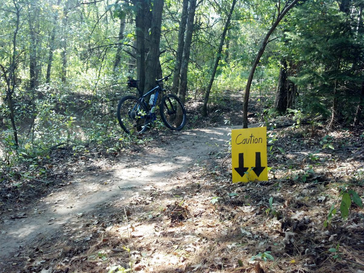 A mountain bike parked beside a dirt path in a wooded area, with a yellow caution sign featuring arrows pointing downwards. The scene is surrounded by greenery, including trees and brush, indicating a natural trail environment. Lowes Creek mountain bike trail.