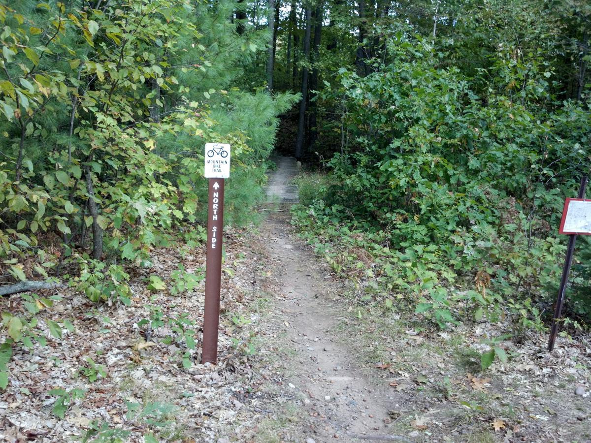 A narrow dirt path leading into a wooded area, flanked by green vegetation and trees. A brown sign on the left indicates "Mountain Trail" with a bicycle symbol, and points toward the "North Side." Another informational sign is visible on the right side of the path. The ground is covered with scattered leaves. Hickory Ridge mountain bike trail.