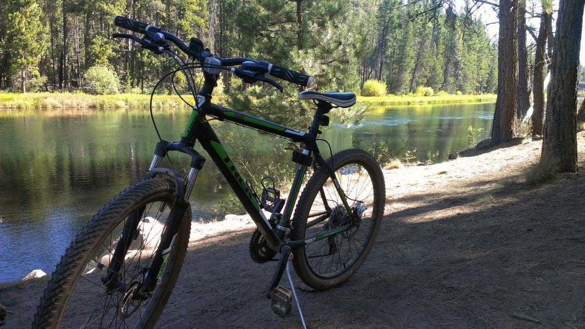 A mountain bike leaning on a stand near a tranquil river, surrounded by a forest of tall trees and green vegetation. The scene captures a peaceful outdoor setting with clear water reflecting the natural surroundings. Deschutes River mountain bike trail.