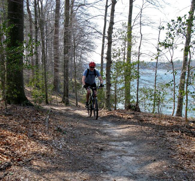 A person riding a mountain bike along a dirt trail bordered by trees, with a body of water visible in the background. The setting is a sunny day with sparse leaves on the trees, suggesting early spring. Beaverdam Park mountain bike trail.