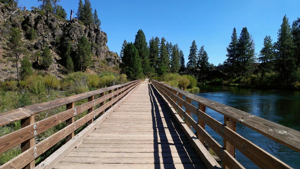 A wooden bridge extends over a calm river, surrounded by lush green trees and rocky hills under a clear blue sky. The perspective is from the beginning of the bridge, leading into the natural landscape. Deschutes River mountain bike trail.