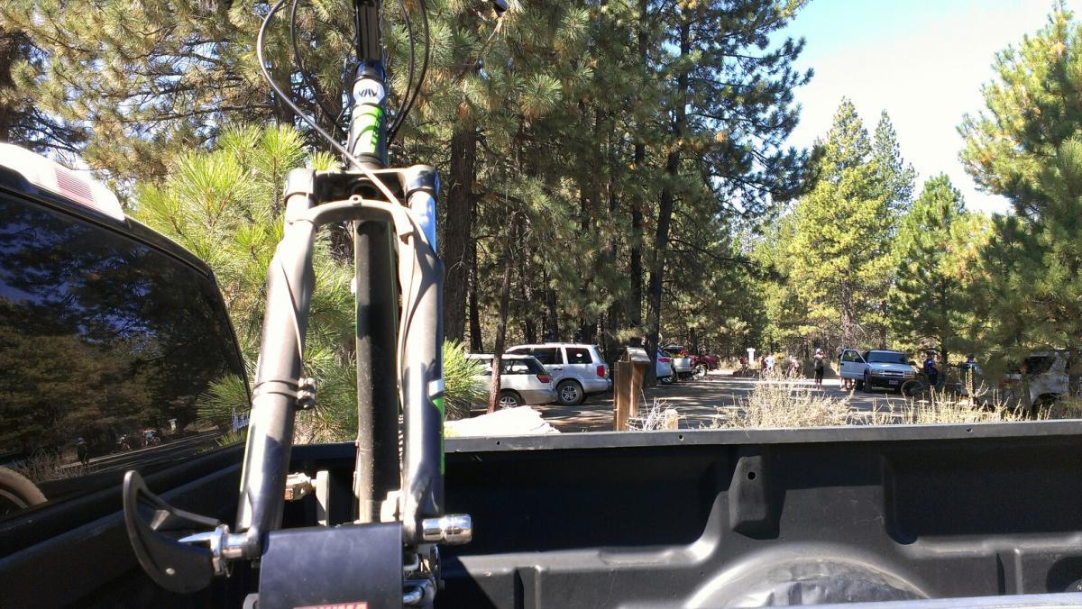 A view from the back of a truck capturing a bicycle frame and handlebars in the foreground, with trees and a parking area visible in the background. Several vehicles are parked along the road, surrounded by a natural landscape of pine trees under a clear blue sky. Phil's Area mountain bike trail.