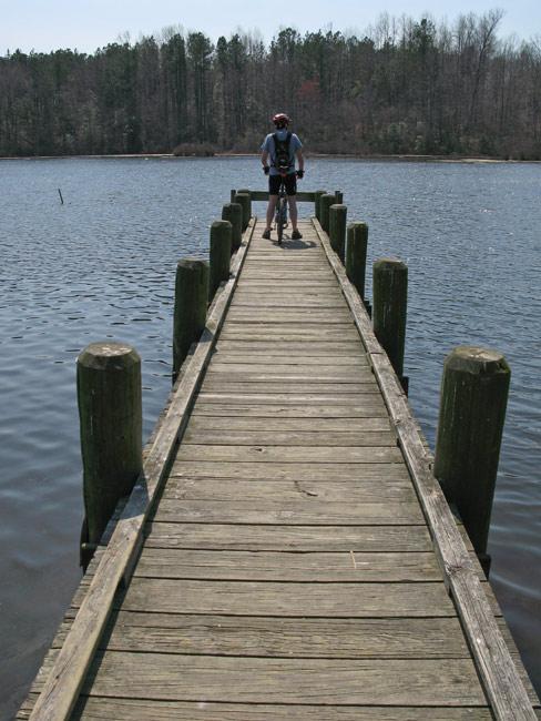 A person standing on a wooden dock that extends into a calm body of water, surrounded by trees in the background on a clear day. Beaverdam Park mountain bike trail.