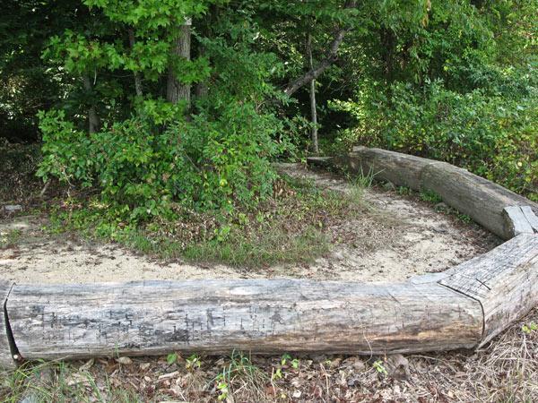 A curved log arc set in a sandy area, surrounded by lush greenery and trees. The log structure appears to be part of a natural landscape, possibly serving as a seating area or boundary in a park or wooded setting. Rosaryville State Park mountain bike trail.