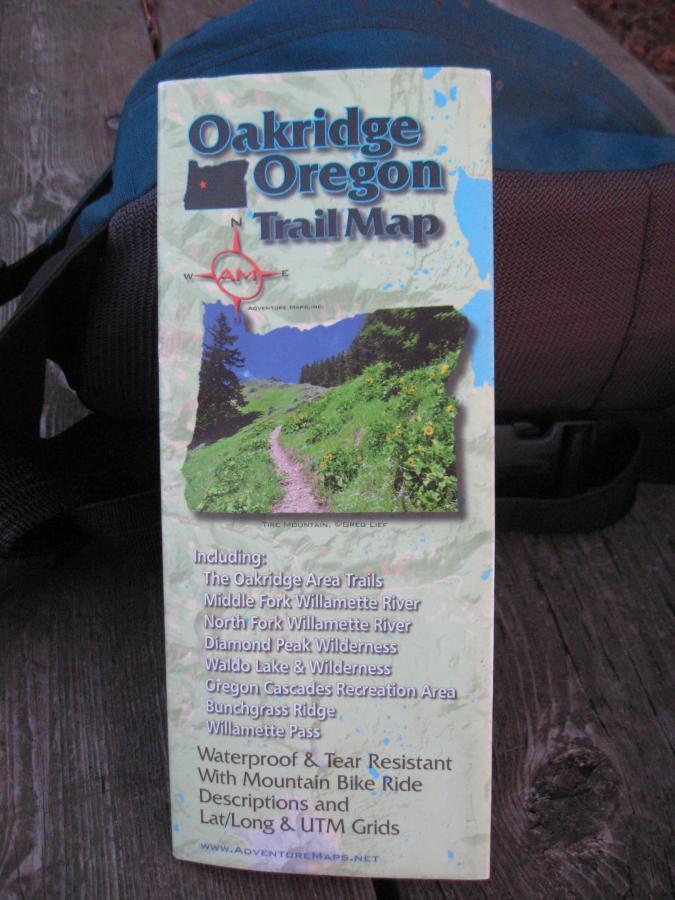 A trail map for Oakridge, Oregon, featuring a scenic pathway surrounded by trees and flowers. The map highlights various areas including the Oakridge Area Trails, Middle Fork Willamette River, Diamond Peak Wilderness, and more. It is labeled as waterproof and tear-resistant, with a compass rose and features descriptions for mountain bike rides. Eula Ridge mountain bike trail.
