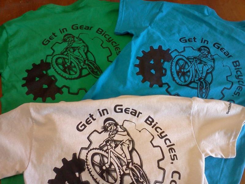 Three t-shirts are displayed, each featuring a graphic design. The shirts are in green, turquoise, and white, with black designs that include the text "Get in Gear Bicycles." The design depicts a cyclist riding a mountain bike inside a gear shape, emphasizing a biking theme.