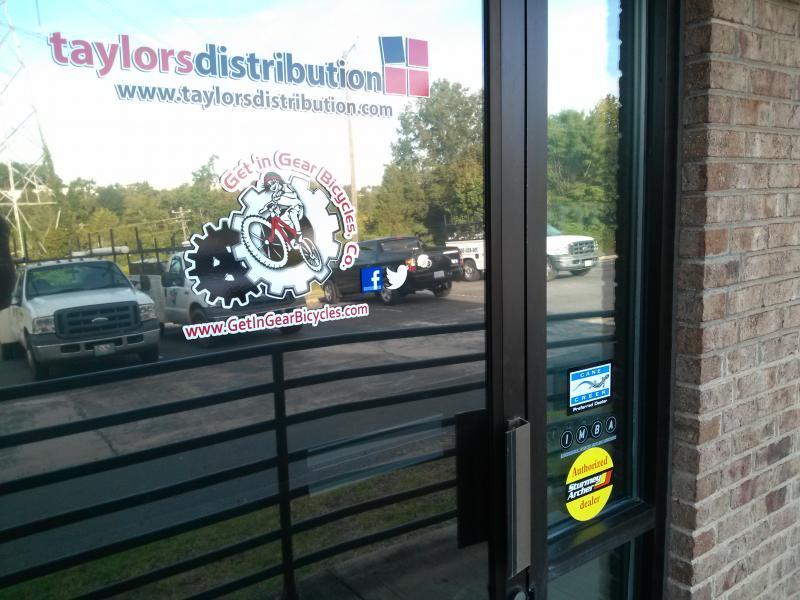 A close-up view of a glass door displaying two business advertisements: one for "Taylors Distribution" with a website link and another for "Get in Gear Bicycles," featuring a logo with a cyclist. The background shows parked vehicles and greenery. A sticker indicating "Authorized Dealer" is also visible.