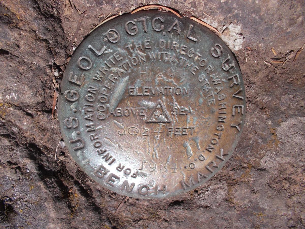 Bronze U.S. Geological Survey benchmark plaque embedded in rock, displaying text that indicates elevation and other geological survey information. The plaque shows an elevation of 3027 feet above sea level, with a date of 1984. Surrounding the plaque, there are rocky textures and some soil. Goodman Creek mountain bike trail.