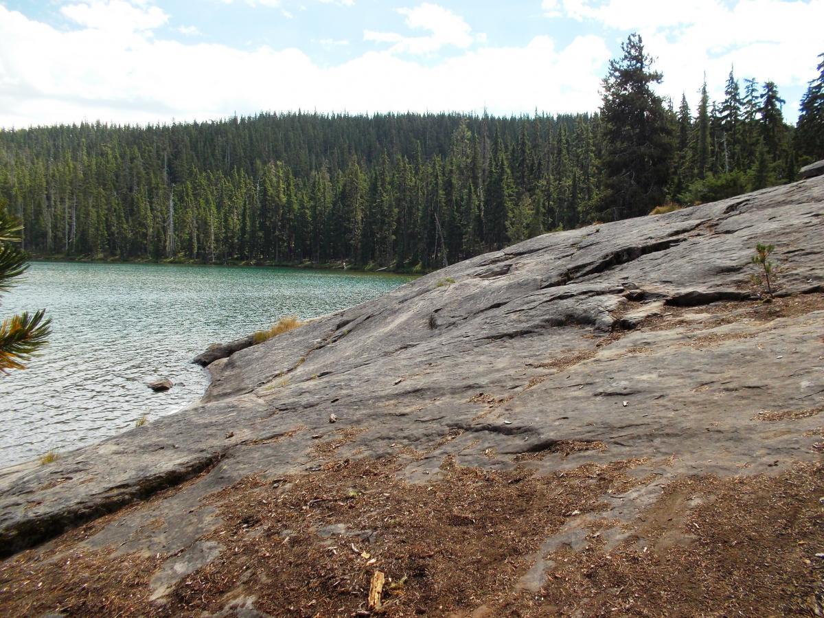 A rocky shoreline beside a clear, turquoise lake, with dense evergreen trees in the background under a partly cloudy sky. Gold Lake To Bobby Lake mountain bike trail.