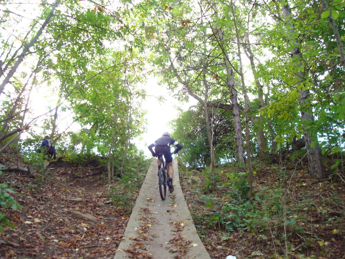 A cyclist riding up a steep, concrete trail surrounded by dense trees in a wooded area. The path is flanked by fallen leaves, and another individual is visible on the left side, climbing the hill. The scene is bright, indicating daylight filtering through the tree canopy above. Frick Park mountain bike trail.