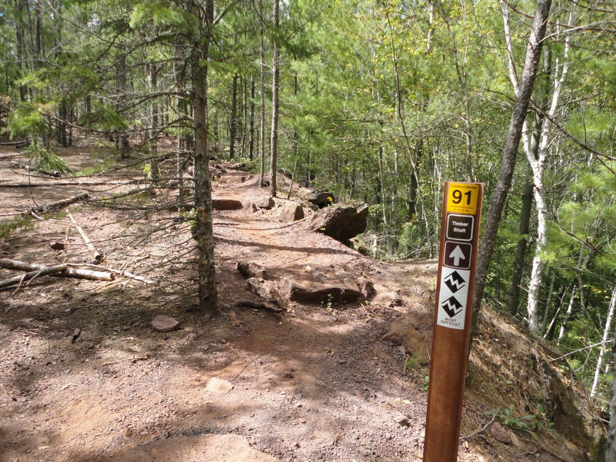 A wooded trail with a dirt path and rocky edges, featuring a trail marker labeled "91" indicating the "Timber Shaft" route. The marker includes directional arrows and symbols indicating "most difficult" terrain, surrounded by tall trees and scattered branches on the ground. Cuyuna Lakes mountain bike trail.