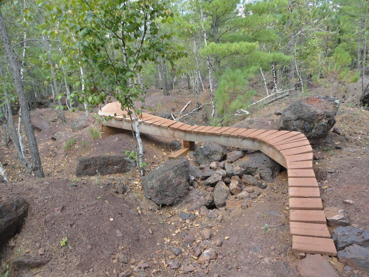 A winding wooden boardwalk meanders through a forested area, surrounded by rocky terrain and scattered trees. The pathway, elevated above the ground, is designed to navigate through natural obstacles, creating a peaceful trail in a lush, green landscape. Cuyuna Lakes mountain bike trail.