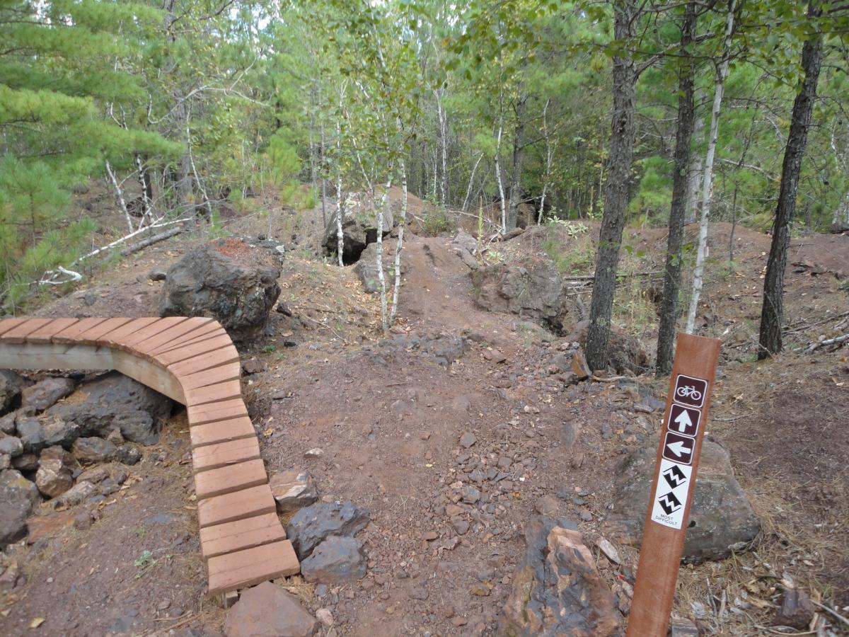 A forested trail featuring a wooden boardwalk that curves to the right, leading into a dirt path surrounded by trees and rocks. A trail sign next to the path indicates directions for biking, with symbols for straight, left, and winding paths. Cuyuna Lakes mountain bike trail.