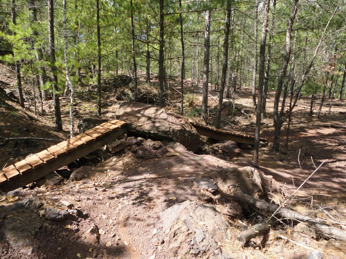 A wooden footbridge spans a small ravine in a forested area, surrounded by tall pine trees and rocky terrain. The trail leading to the bridge is unpaved, with scattered leaves and stones. Sunlight filters through the tree canopy, creating dappled light on the ground. Cuyuna Lakes mountain bike trail.