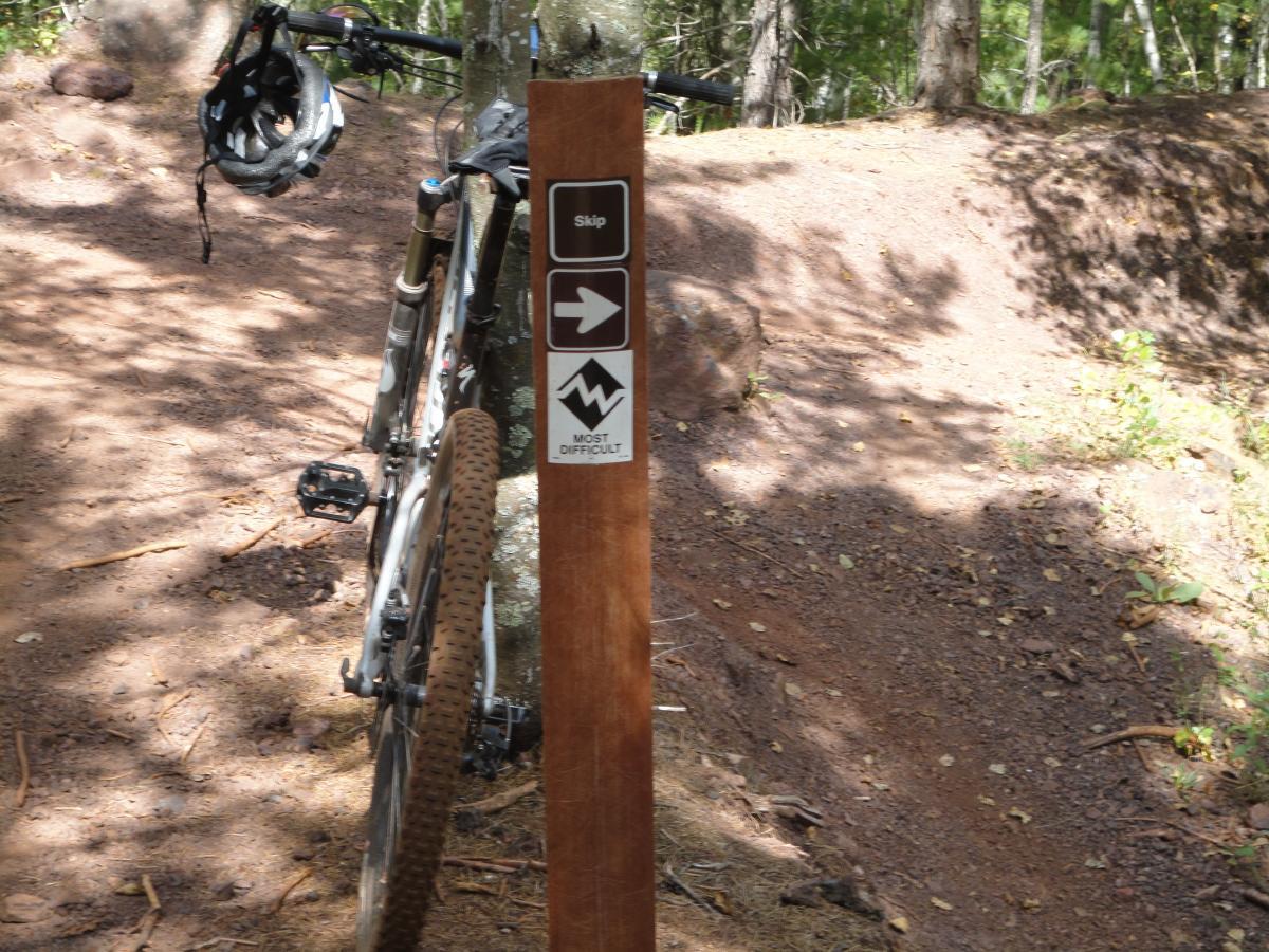 A mountain bike parked beside a trail sign marking a difficult route. The sign features indicators for "Skip" and "Most Difficult" along with directional arrows. The surrounding area is a wooded path with a dirt surface. Cuyuna Lakes mountain bike trail.