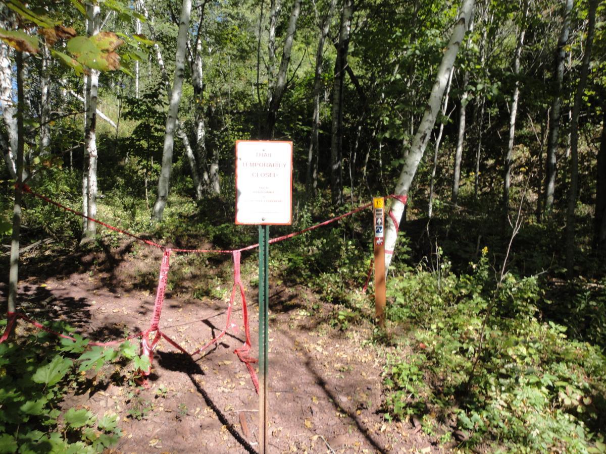 A pathway in a forest environment, marked with a "Trail Temporarily Closed" sign on a post. Red tape is used to block off the entry to the trail, and the surrounding area features various trees and underbrush, indicating a natural outdoor setting. Cuyuna Lakes mountain bike trail.