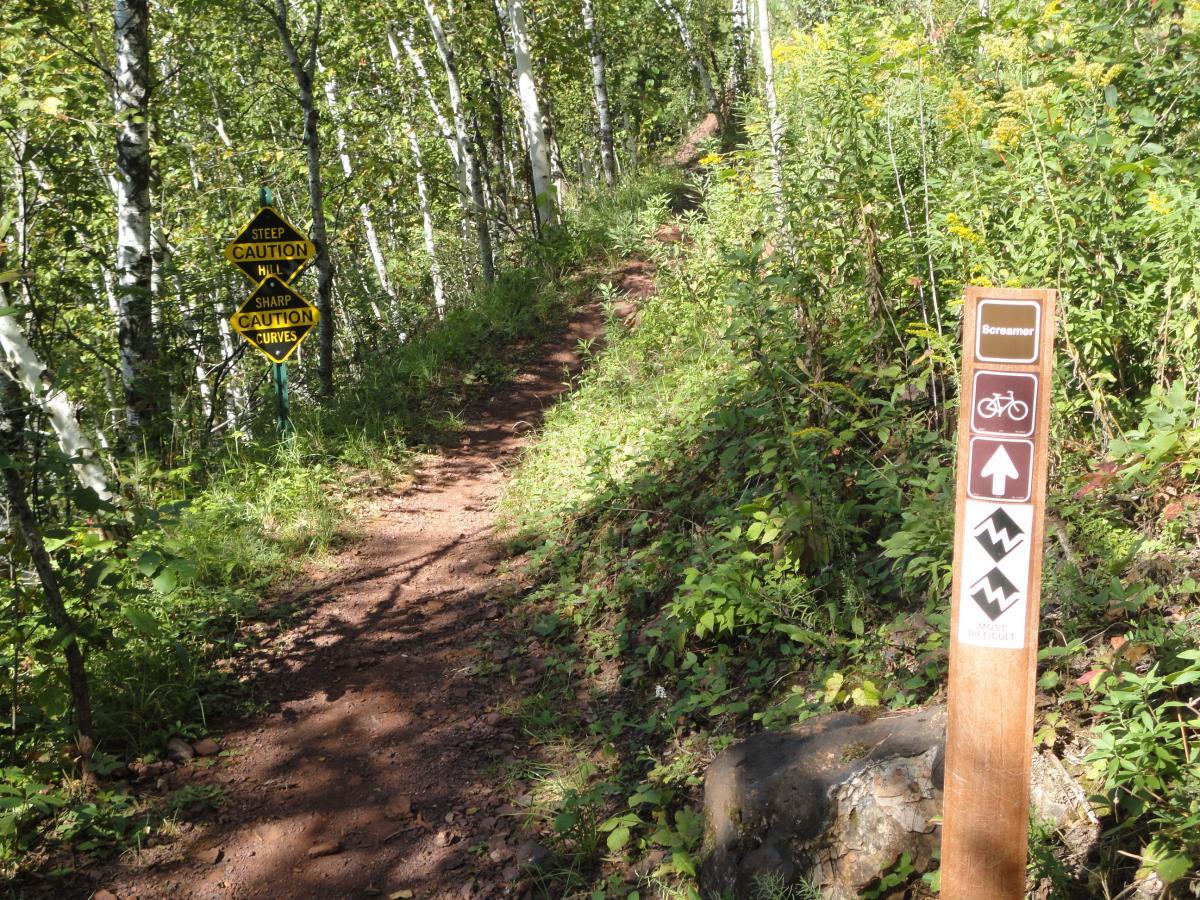 A winding dirt trail surrounded by greenery and trees, featuring warning signs for steep hills and sharp curves. A wooden post with trail difficulty indicators and a bike symbol stands on the side of the path, inviting outdoor enthusiasts to explore the area. Cuyuna Lakes mountain bike trail.