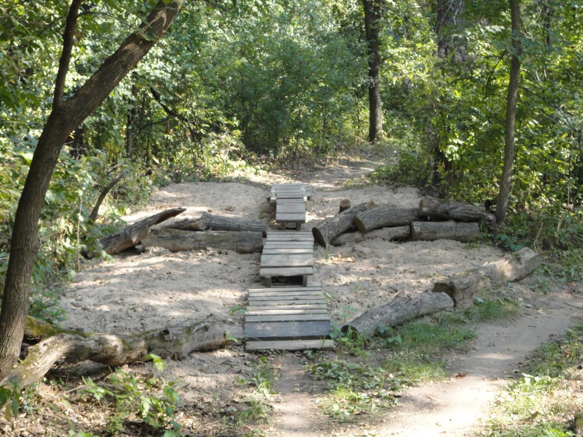 A wooden bridge made from planks crosses a sandy area surrounded by greenery in a forested setting. Logs are arranged on either side of the bridge, adding to the natural landscape. Hillside Park mountain bike trail.